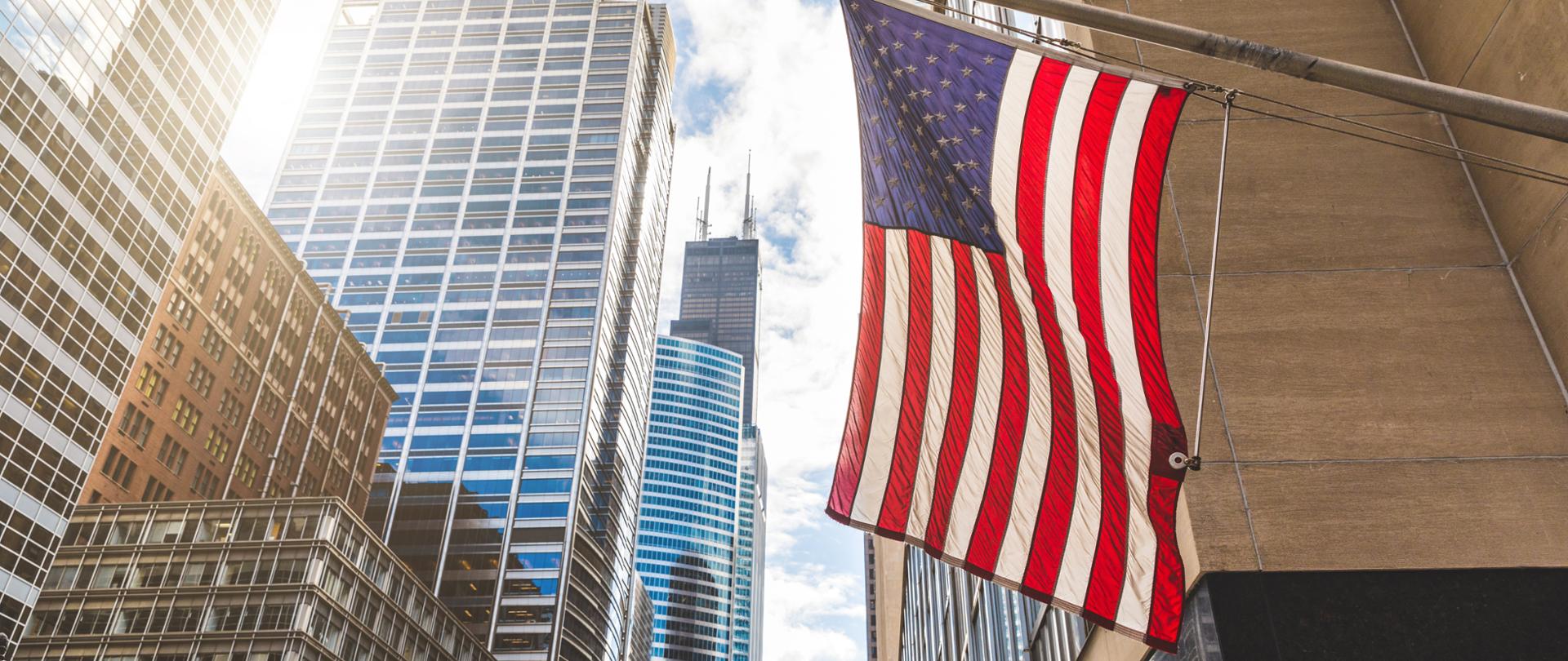 USA flag in Chicago with with skyscrapers on background. American flag waving in the city on a sunny day. Clouds reflections on buildings facade