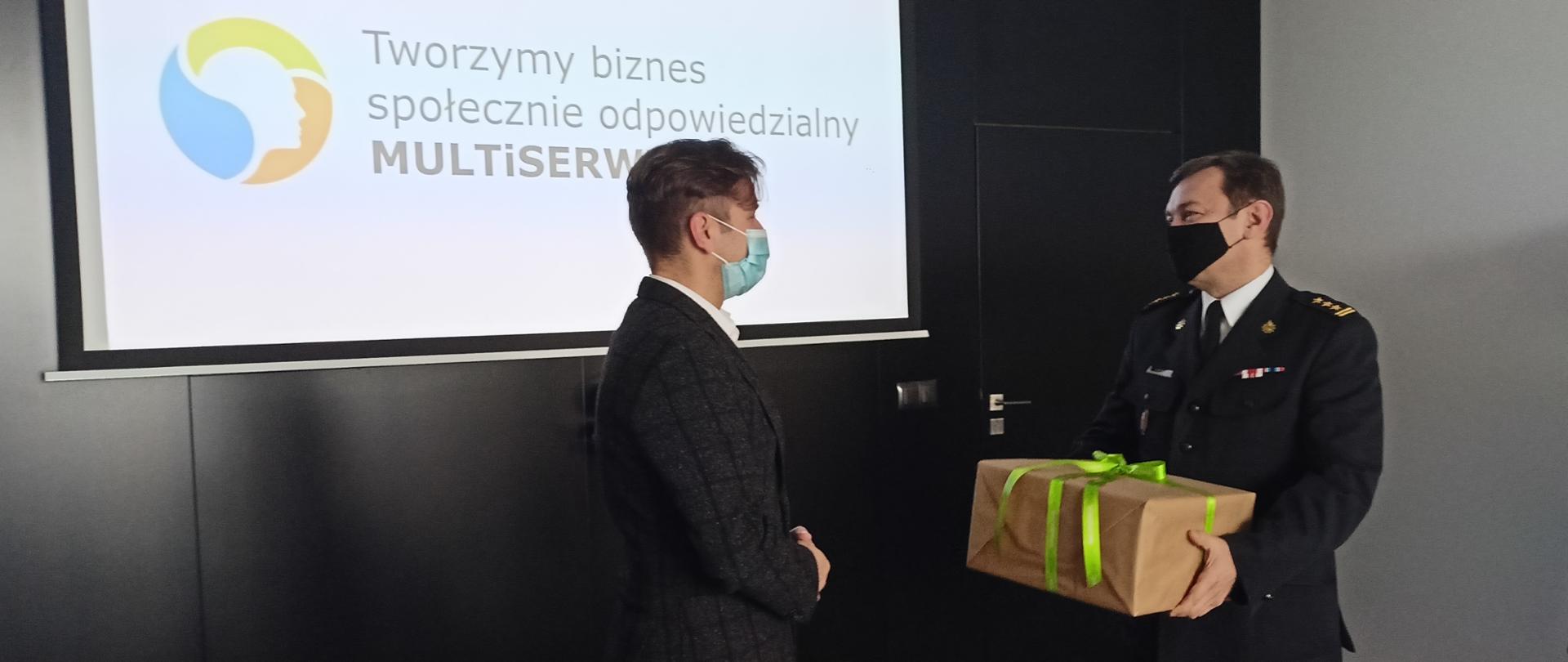 Na czarnej ścianie rozwinięty ekran z wyświetlonym slajdem z napisem: „Tworzymy biznes społecznie odpowiedzialny MULTISERWIS. Na tle tej ściany , na środku planu, stoi zwrócony do patrzącego prawym bokiem prezes firmy Multiserwis ubrany w ciemnoszare spodnie i marynarkę szarą w czarną dużą kratę. Po prawej stronie Komendant Powiatowy PSP w Krapkowicach, w mundurze wyjściowym trzymający w rękach zapakowany w papier do pakowania i związany zieloną wstążką „prezent”. Drugi, mniejszy prezent leży na stole na pierwszym planie. 
