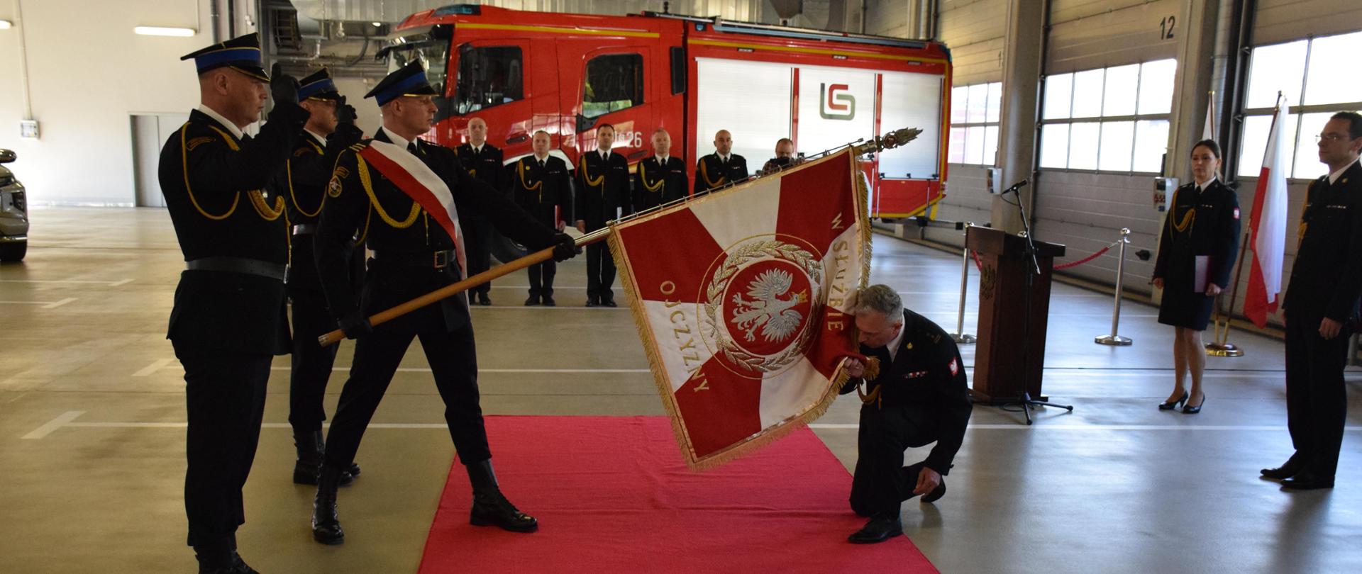 Nowo powołany Komendant Miejski PSP w Mysłowicach klęczy na czerwonym dywanie i całuje sztandar JRG Mysłowice trzymany przez poczet sztandarowy