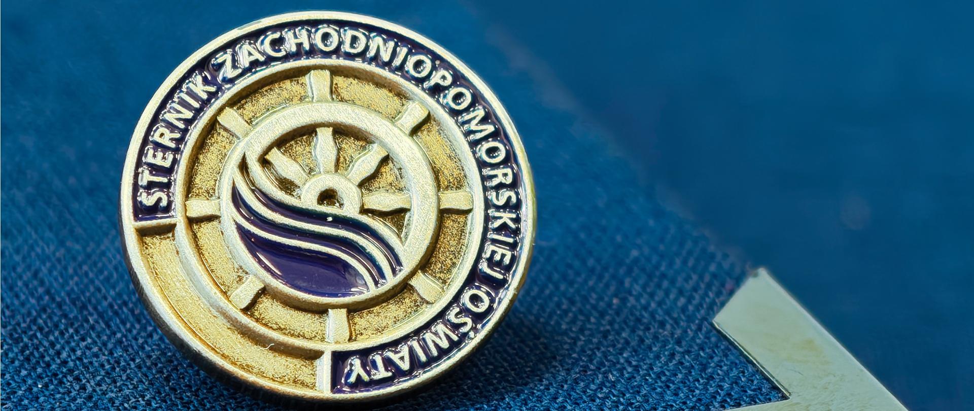 na niebieskim tle srebrna odznaka Sternik Zachodniopomorskiej Oświaty 