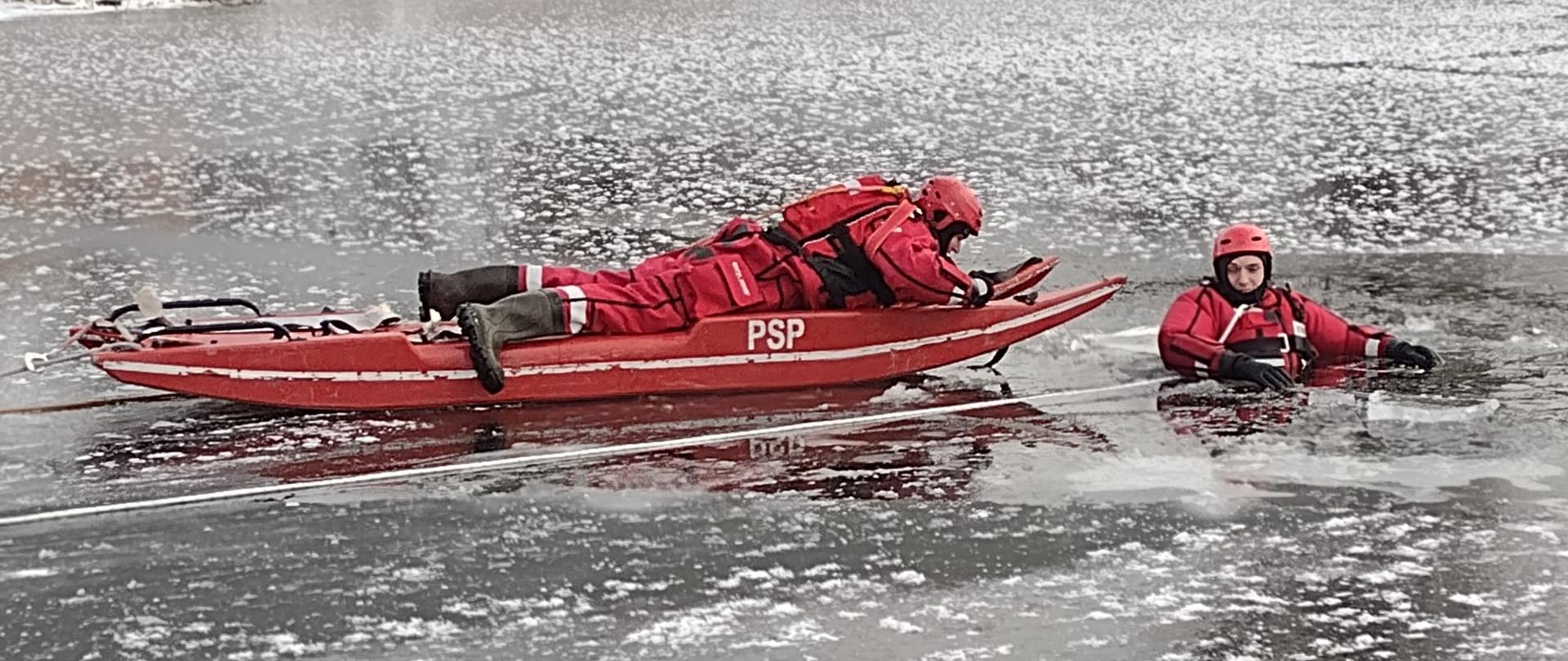 Dwóch strażaków znajduje się z dala od brzegu. Jeden ze strażaków znajduje się w wodzie. Drugi strażak znajduje się w bliskiej odległości w pozycji leżącej na deskosaniach.
Strażacy ubrani w kombinezony wypornościowe koloru czerwonego, zabezpieczeni linkami ratowniczymi.