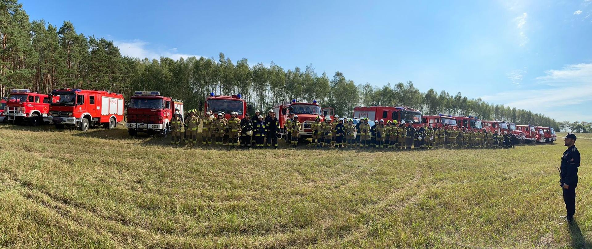 Fotografia panoramiczna przedstawiająca pojazdy kompanii gaśniczej, a przed nimi ustawionych kilkudziesięciu strażaków, stojących na polanie.