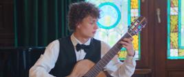Zdjęcie przedstawia młodego chłopaka w koszuli, muszce i kamizelce grającego na gitarze. Jedną stopę trzyma na podstawce. Siedzi na krześle. W tle widać okno z witrażami i zielone zasłony.