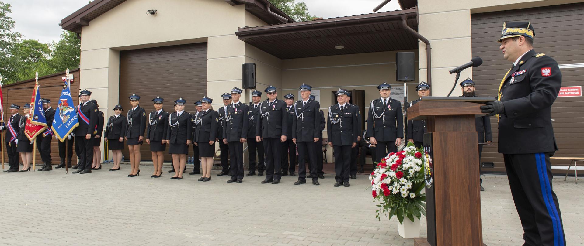 Panorama uroczystego apelu z okazji 100-lecia OSP w Wilkowie Polskim przed strażnicą. Na pierwszym planie Mazowiecki Komendant Wojewódzki PSP nadbryg. Artur Gonera przemawia przy mównicy, a w tle stoją strażacy, poczty sztandarowe oraz uczestnicy uroczystości