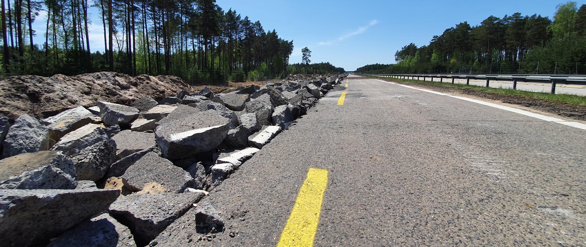 Na zdjęciu widać drogę krajową nr 18. Z lewej strony rozkruszone na duże kawałki stare płyty jedni betonowej prawego pasa jezdni południowej. Lewy pas jeszcze w całości. Za barierami z prawej strony zdjęcia widać północną jezdnie drogi. Po obu stronach las.
