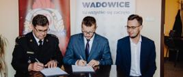 Widok z przodu. Moment podpisywania porozumienia w sprawie przekazania sprzętu. Za stołem od lewej siedzą: Komendant Powiatowy PSP w Wadowicach st. bryg. Paweł Kwarciak, Burmistrz Wadowic Bartosz Kaliński, Poseł na Sejm RP Filip Kaczyński. Komendant i Burmistrz podpisują dokumenty. 