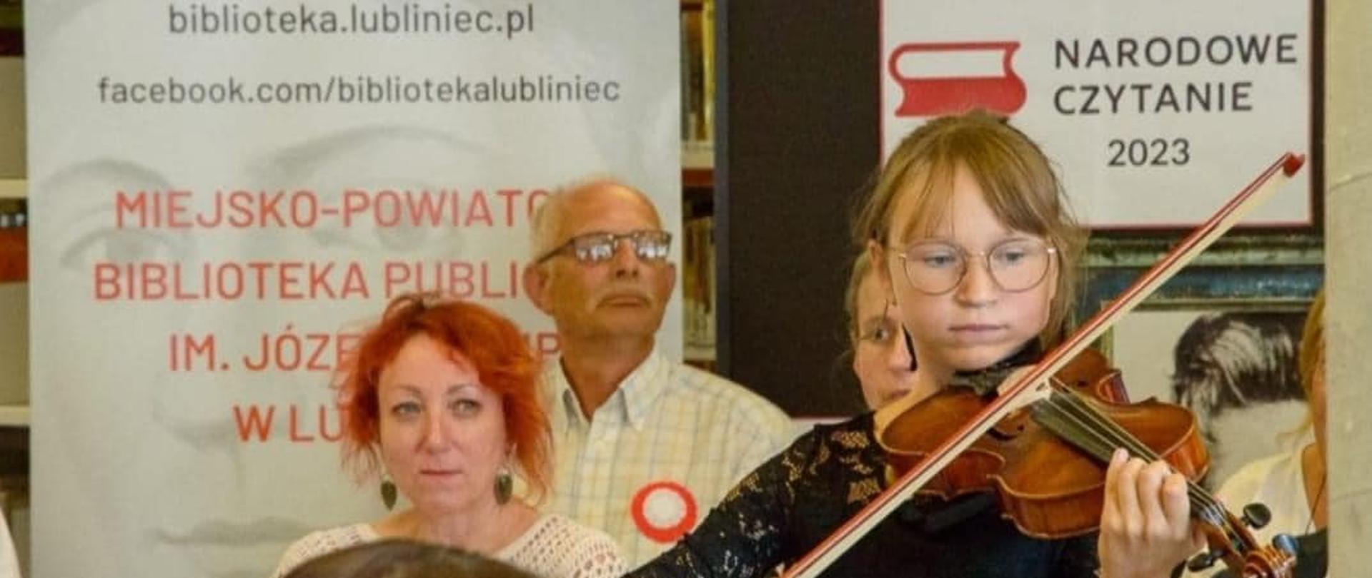 Dziewczynka w ciemnym ubraniu gra na skrzypcach. W tle plakat z akcji Narodowe Czytanie, baner Lubiteki oraz kilkoro ludzi zgromadzonych na tym wydarzeniu
