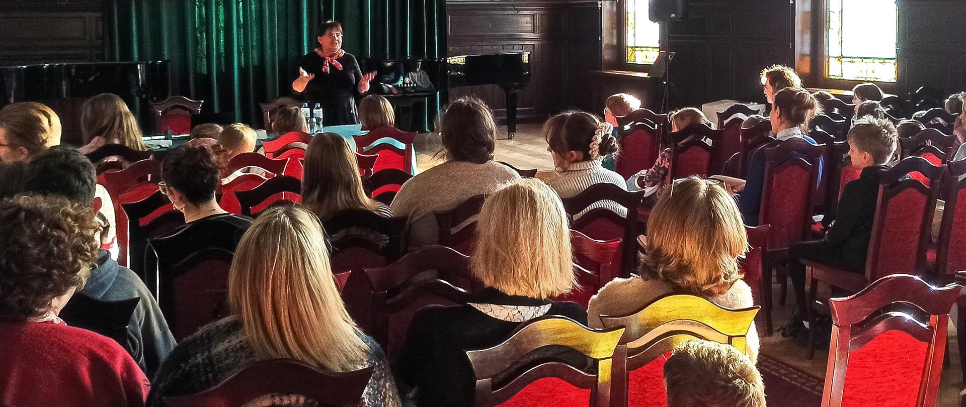 Zdjęcie podczas wykładu . Na czerwonych krzesłach siedzi publiczność, która ogląda przemawiającą na scenie kobietę. Jest ona ubrana na czarno, a na szyi przewiązana ma czerwoną apaszkę. Za nią znajduje się fragment ekranu z prezentacją multimedialną, fortepian oraz długa zielona zasłona.
