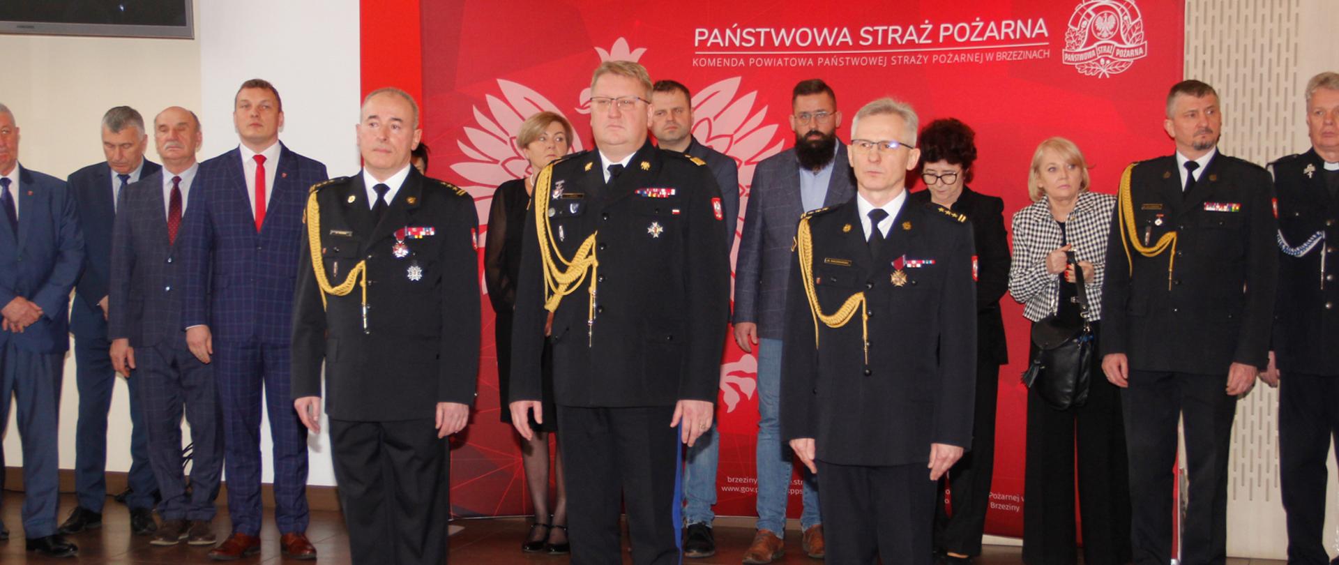 trzech strażaków stojących w szeregu za nimi inni uczestniczy uroczystości