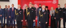 trzech strażaków stojących w szeregu za nimi inni uczestniczy uroczystości
