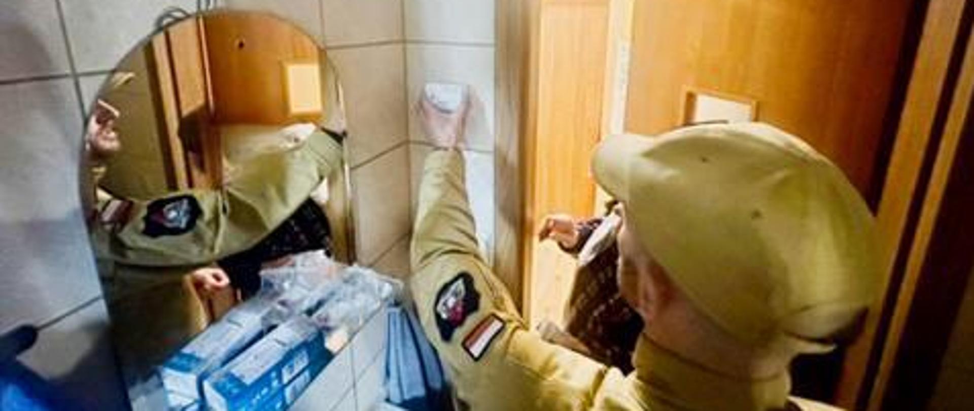 Strażak w beżowym mundurze montuje czujnik tlenku węgla na ścianie w łazience. W lustrze odbija się wnętrze pomieszczenia oraz stojący obok mieszkaniec.