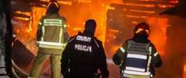 Na zdjęciu w porze nocnej widoczni dwaj strażacy w środku policjant. W oddalali objęty pożarem dom.
