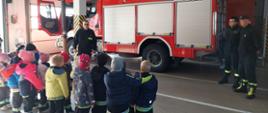 Zdjęcie przedstawia dzieci z przedszkola które stoją w garażu jednostki ratowniczo - gaśniczej w Koninie. Strażacy zapoznają dzieci z pracą strażaka. Z tyłu widać samochód strażacki. 