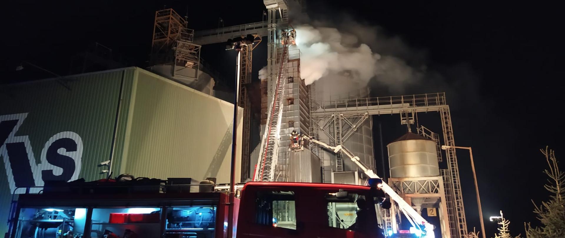 Zdjęcie wykonane w nocy. Na pierwszym planie bokiem do widza stoi samochód ratowniczo-gaśniczy koloru czerwonego marki Scania (numer operacyjny 402o25) z otwartymi skrytkami przestrzeni roboczej oświetlonych światłem led w każdej z nich. Na drugim planie znajduje się część zakładu wraz suszarnią (pionowy część wysokości kilkunastu metrów) z której wydobywa się (górna część) jasny gęsty dym. Obiekt zakładu oświetlony sztucznym światłem dobrze widoczny. Na zdjęciu widoczna część drabiny mechanicznej w koszu której znajdują się strażacy na wysokości wydostającego się dymu z suszarni. W prawej części kadru znajduje się podnośnik mechaniczny w koszu którego znajdują się strażacy. Podnośnik rozwinięty do połowy wysokości suszarni.