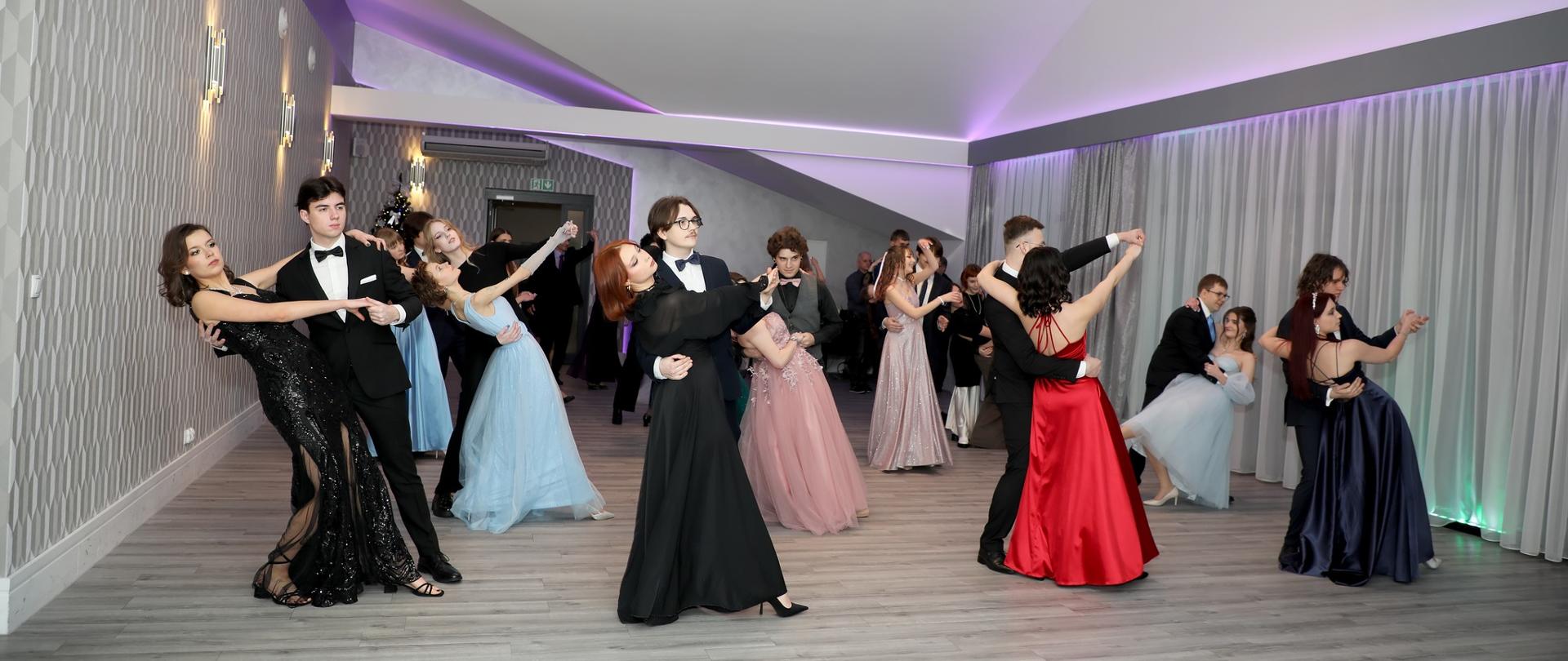 Zdjęcie przedstawia grupę osób w strojach wieczorowych, które tańczą w jasnym, nowoczesnym pomieszczeniu. Osoby są sparowane i wydają się być zaangażowane w taniec. 