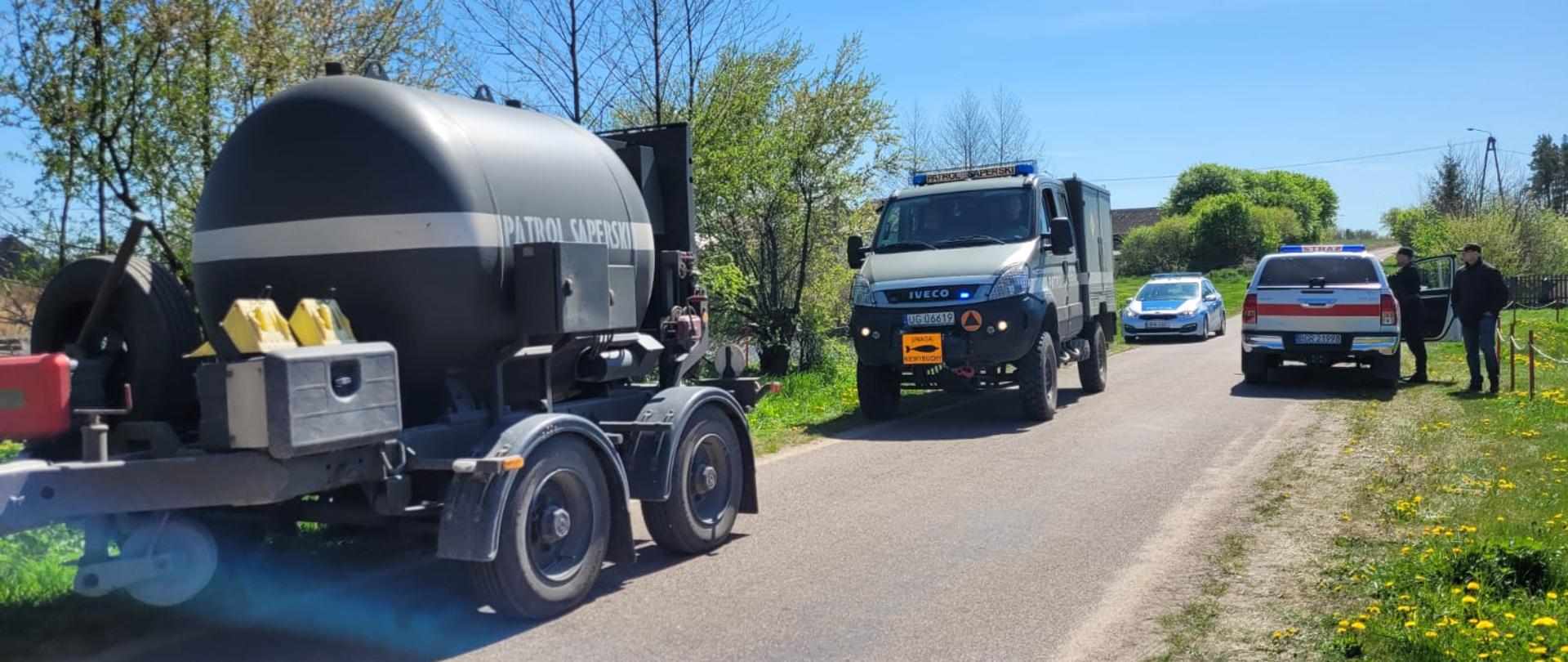 Pojazdy patrolu saperskiego oraz policji i straży pożarnej stoją na drodze.