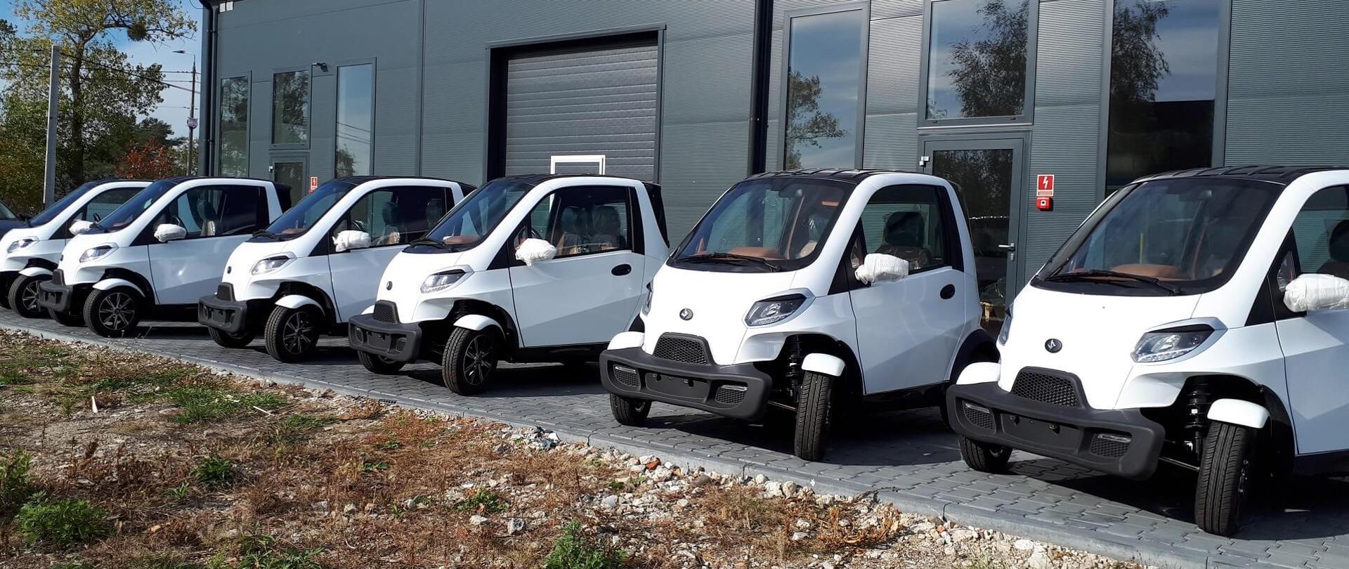 Rząd sześciu biało-czarnych aut przed halą przemysłową