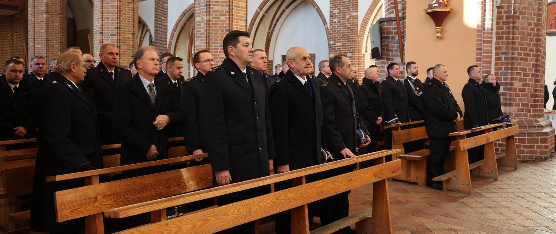 Na zdjęciu widać grupę mężczyzn stojących w kościele.
Mężczyźni stoją w ławkach i patrzą w jednym kierunku. Większość z nich ma na sobie ciemne, eleganckie mundury. Kościół ma jasne ściany i dużo cegieł. Widać także złotą figurę na ścianie i duże łuki z cegły.