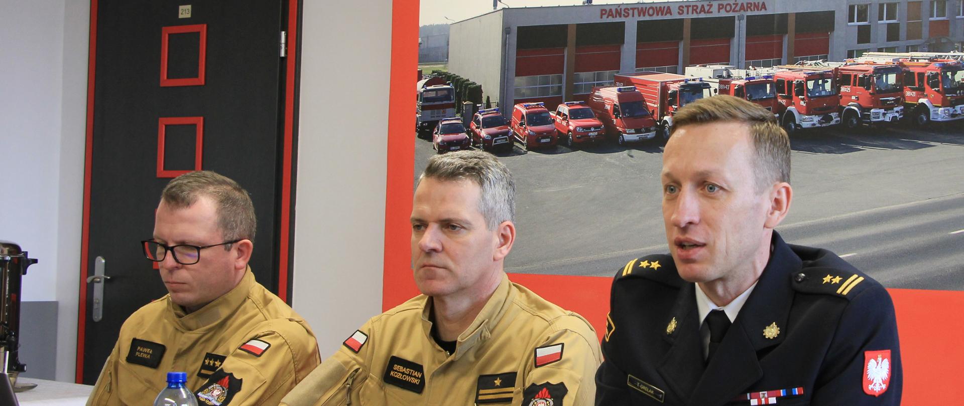 trzech strażaków siedzi przy stole, za nimi slajd z duża ilością pojazdów 