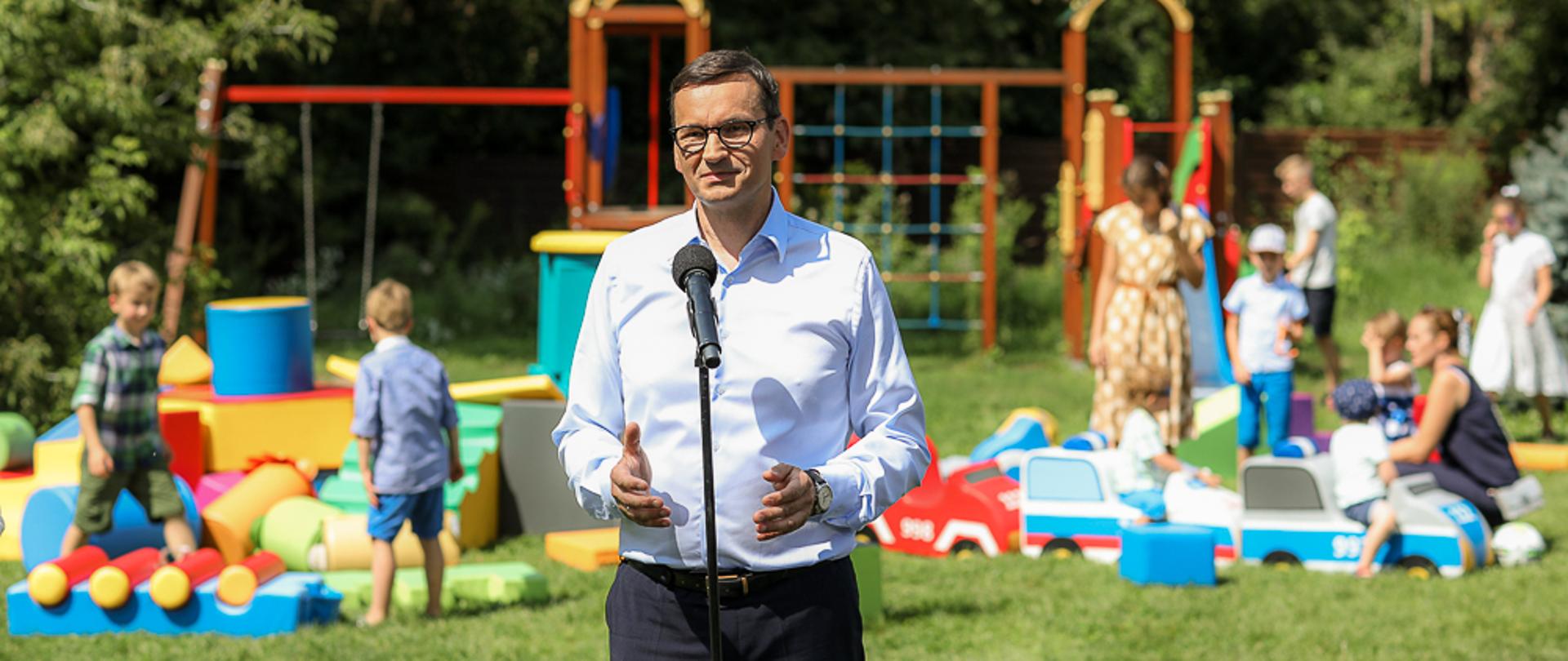 Premier Mateusz Morawiecki stoi przed mikrofonem, w tle plac zabaw, na którym bawią się dzieci,