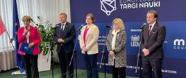 Zdjęcie z boku pod kątem, w pomieszczeniu z zieloną podłogą przy mikrofonach na stojakach stoi pięć osób, pośrodku wiceminister Zioło-Pużuk, za nimi przed zasłoniętym białymi zasłonami oknem baner z napisem Europejskie Targi Nauki.