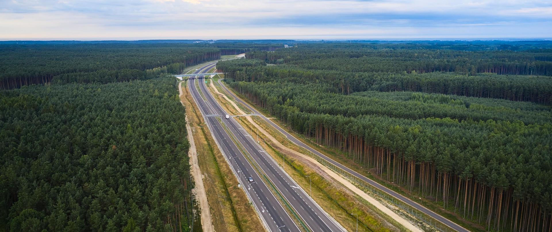 Widok z góry na dwujezdniową drogę ekspresową. Na środku biegnie droga, po której jadą auta. Po bokach lasy. W dalszej perspektywie widać wiadukt nad drogą ekspresową. 