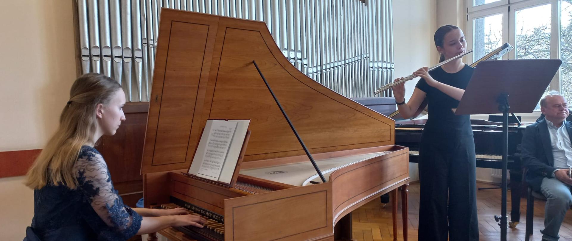 na zdjęciu na pierwszym planie dziewczyna w blond włosach i granatowej sukni grająca na klawesynie na drugim planie dziewczyna w ciemnych włosach spiętych w kitkę i czarnym ubrani grająca na flecie przed nią pulpit na nuty