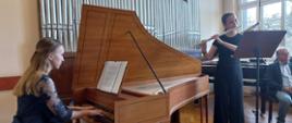 na zdjęciu na pierwszym planie dziewczyna w blond włosach i granatowej sukni grająca na fortepianie na drugim planie dziewczyna w ciemnych włosach spiętych w kitkę i czarnym ubrani grająca na flecie przed nią pulpit na nuty