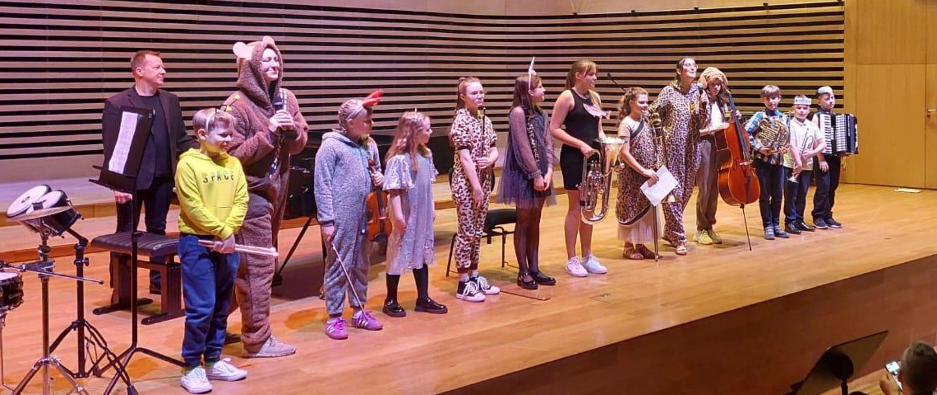 Wszyscy wykonawcy - jedenaścioro dzieci z instrumentami i dwie kobiety w przebraniach różnych zwierzątek oraz mężczyzna stoją w rzędzie na estradzie sali koncertowej.