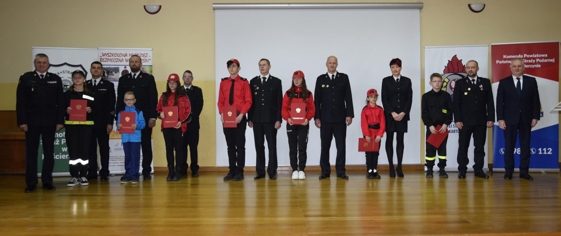 Wspólne zdjęcie strażaków zaproszonych gości oraz przedstawicieli Młodzieżowych Drużyn Pożarniczych którzy trzymają czerwone teczki.