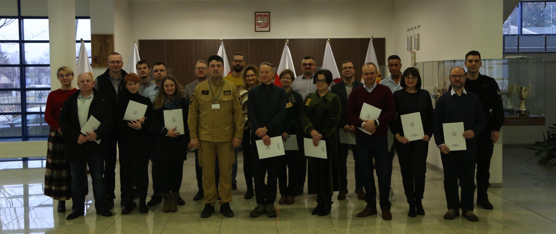 Grupowe zdjęcie uczestników szkolenia (19 osób) w towarzystwie Komendanta Centralnej Szkoły PSP oraz oficera CS PSP. Uczestnicy szkolenia trzymają otrzymane zaświadczenia