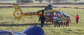 Zdjęcie przedstawia strażaków, ratowników medycznych oraz policjanta. Strażacy przekazują osobę poszkodowaną do śmigłowca Lotniczego Pogotowia Ratunkowego. Śmigłowiec stoi na polu.