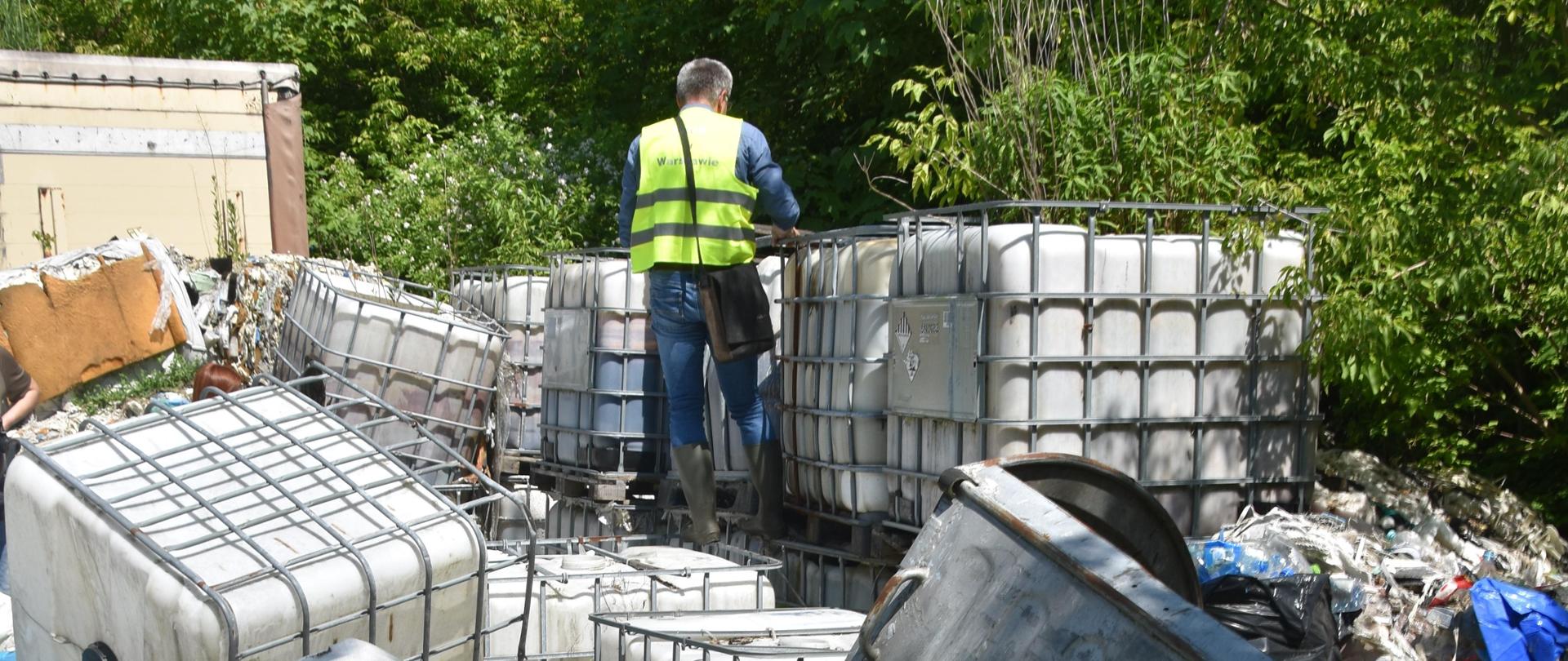 Inspektor Wojewódzkiego Inspektoratu Ochrony Środowiska w Warszawie prowadzi oględziny pojemników typu mauzer ustawionych jedne na drugich. W tle widać tył naczepy samochodowej oraz bujną, zieloną roślinność.