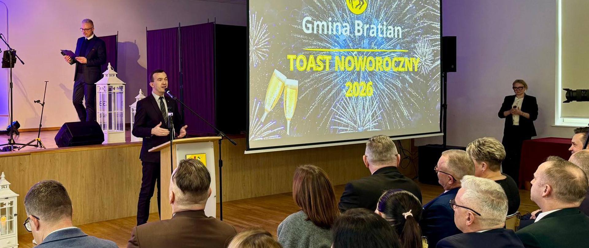 mężczyzna w garniturze przemawia przez mikrofon na mównicy na tle prezentacji "Toast Noworoczny Gmina Bratian"