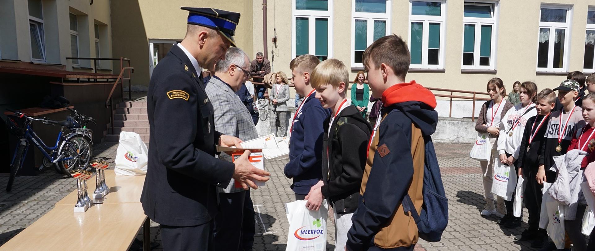 Wręczanie nagród dla zwycięzców indywidualnych w grupie II. Młodzieży nagrody wręcza zastępca komendanta PSP w Grajewie. W tle budynek szkoły.