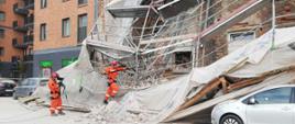 Gruzowisko na ul. Wrzesińskiej. Fragmenty zburzonej śiany kamienicy oraz powalonej konstrukcji rusztowania. Przygnieciony samochód. Dwóch ratowników w pomarańczowych kombinezonach przeszukuje gruzowisko przy użyciu specjalistycznego sprzętu.