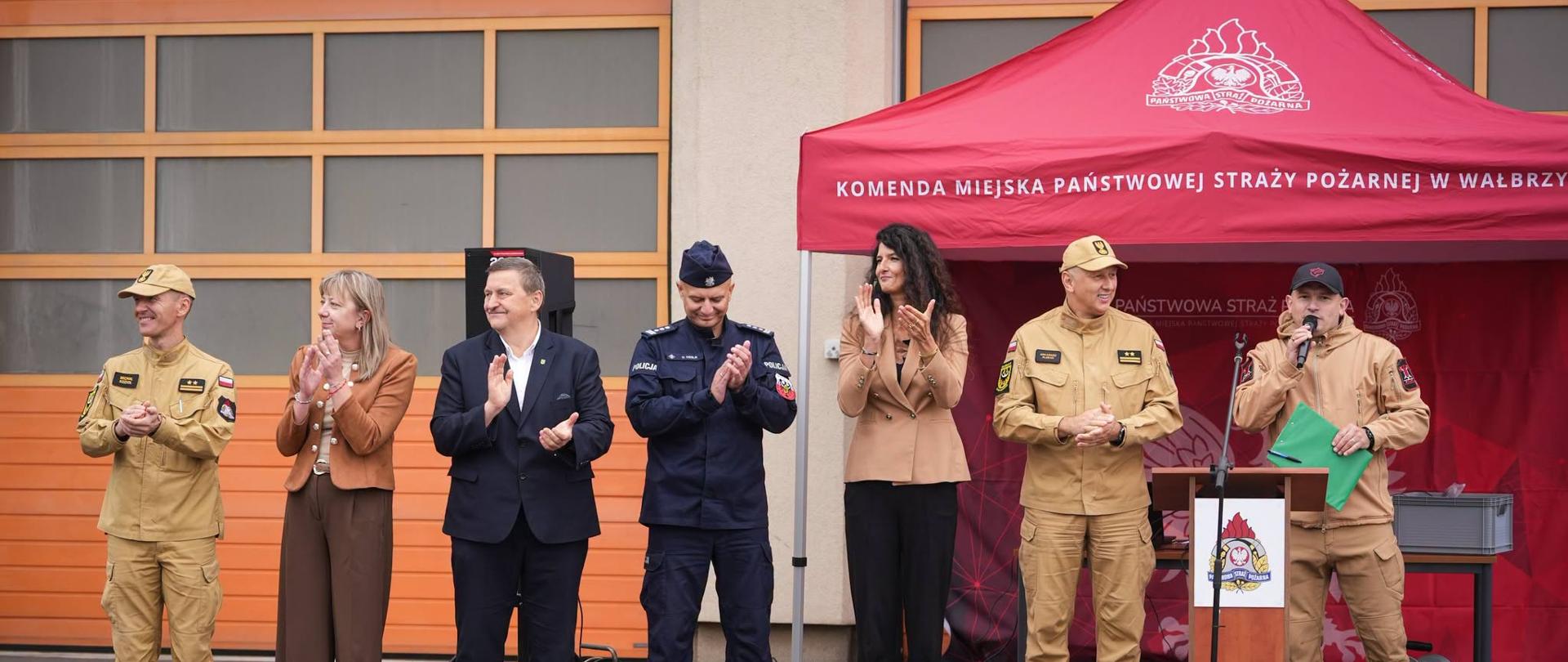 Na zdjęciu sześć osób stoi w rzędzie przed czerwonym namiotem Komendy Miejskiej Państwowej Straży Pożarnej w Wałbrzychu. Grupa składa się z umundurowanych strażaków, policjanta oraz dwojga oficjalnie ubranych cywilów. Jeden ze strażaków stoi przy drewnianej mównicy i przemawia do mikrofonu, trzymając w dłoni zieloną teczkę. Reszta osób uśmiecha się i bije brawo. W tle widać elewację budynku z pomarańczowymi bramami garażowymi