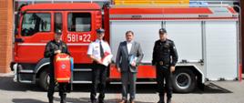 Przekazanie plecaka przez zastępce dyrektora WORD w szarym garniturze w obecności Komendanta Powiatowego oraz zastępcy Komendanta Powiatowego na tle samochodu strażackiego . Plecak trzyma strażak za nim budynek komendy z czerwonej cegły 