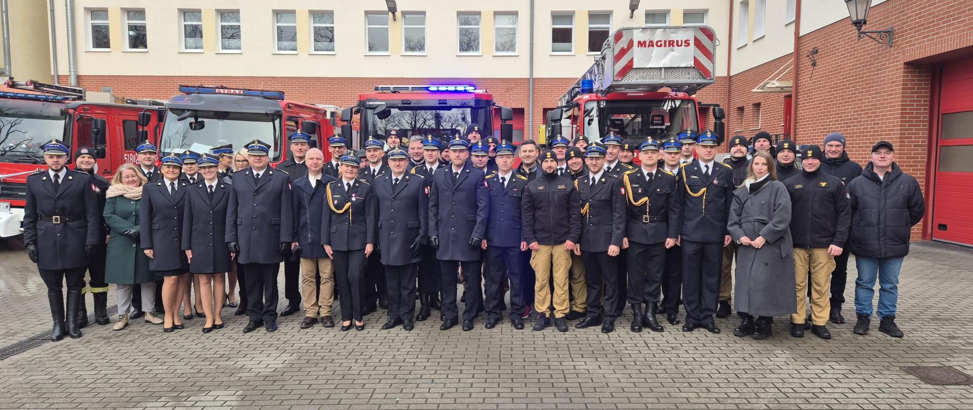 Na zdjęciu widać grupę strażaków na zdjęciu grupowym, stojących przed samochodami gaśniczymi. 