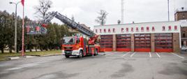 Samochód pożarniczy drabina a w tle drzewa i budynek strażnicy