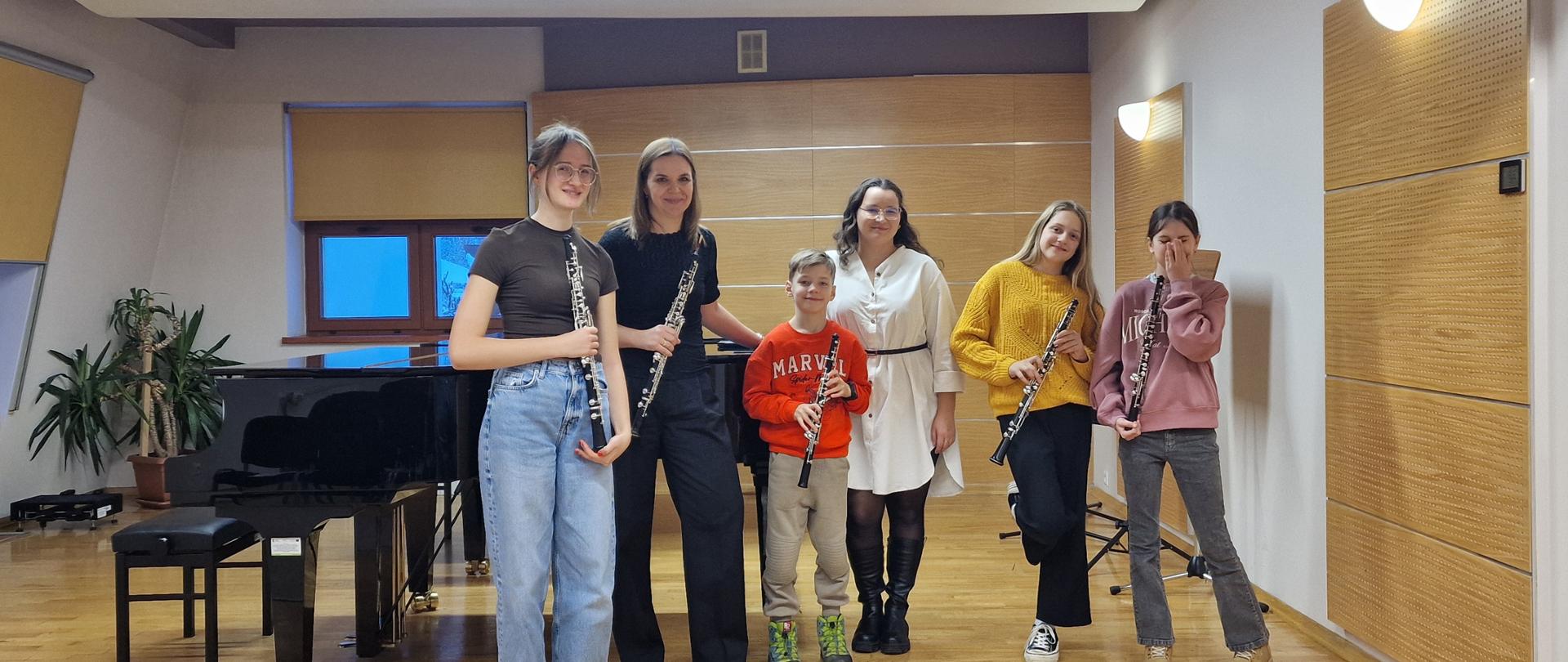 To zdjęcie grupowe zostało wykonane we wnętrzu sali koncertowej lub lekcyjnej, prawdopodobnie w szkole muzycznej. Na drewnianej scenie, na pierwszym planie, stoi sześć osób – dwie dorosłe kobiety i czwórka dzieci/młodzieży, prawdopodobnie z klasy instrumentów dętych drewnianych.Osoby na zdjęciu: Po lewej stronie stoi młoda dziewczyna w jasnych, luźnych dżinsach i ciemnej bluzce z długim rękawem, trzymająca klarnet. Obok niej stoi dorosła kobieta ubrana całkowicie na czarno. Jest wysoka i trzyma klarnet oparty o podłogę. Wygląda na nauczycielkę. W środku stoi mały chłopiec w czerwonym swetrze i szarych spodniach, trzymający w ręku mały klarnet lub inny instrument dęty drewniany. Obok niego stoi druga dorosła kobieta w jasnej, luźnej, długiej bluzce lub sukience, przewiązanej w pasie czarnym paskiem, patrząca w kierunku obiektywu z uśmiechem. Kolejna młoda dziewczyna w żółtym, wełnianym swetrze i ciemnych spodniach również trzyma klarnet. Skrajnie po prawej stoi najmłodsza dziewczynka w różowym/jasnym swetrze i dżinsach, która podnosi dłoń do ust i śmieje się. Również ma klarnet. Tło: W tle, po lewej stronie, widać czarny fortepian z otwartym wiekiem, a za nim okno. Ściany są wyłożone jasnymi, drewnianymi panelami w geometryczne wzory, poprawiającymi akustykę. Sufit ma podłużne, świetlówkowe oświetlenie. Cała scena jest dobrze oświetlona, a nastrój jest radosny i nieformalny. Zdjęcie przedstawia prawdopodobnie mały zespół lub klasę klarnetu po zajęciach lub próbie.