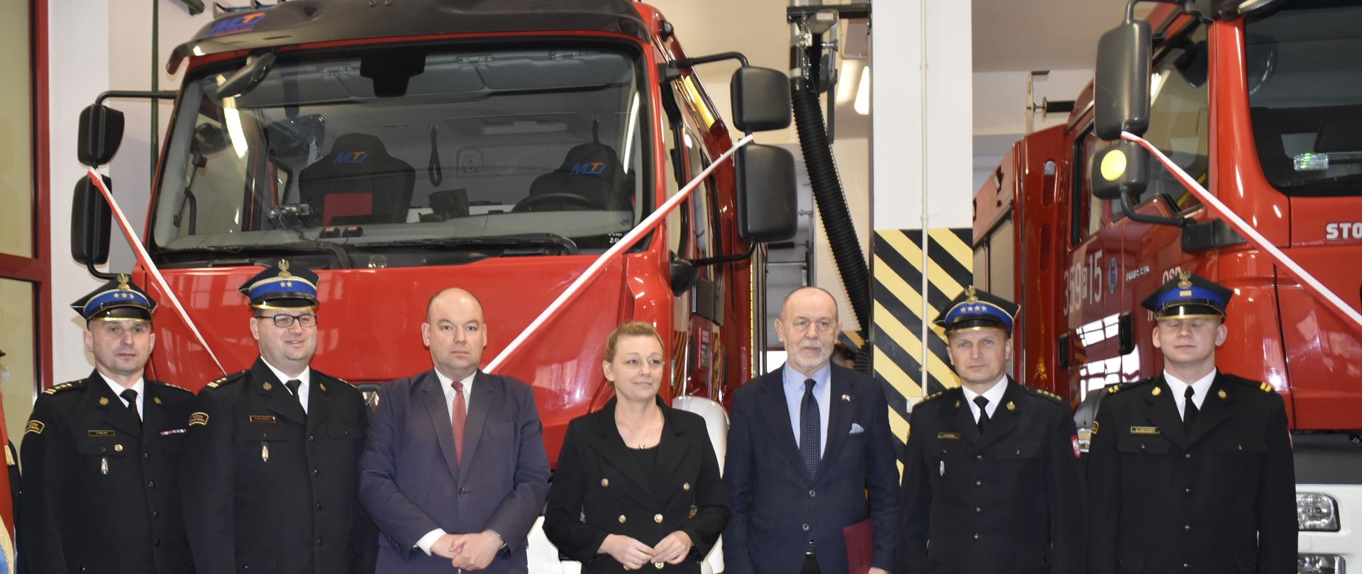 grupa luszi w tym strażacy w rogatywkach stoją przed czerwonym samochodem strażackim z biało czerwoną wstęgą na przodzie