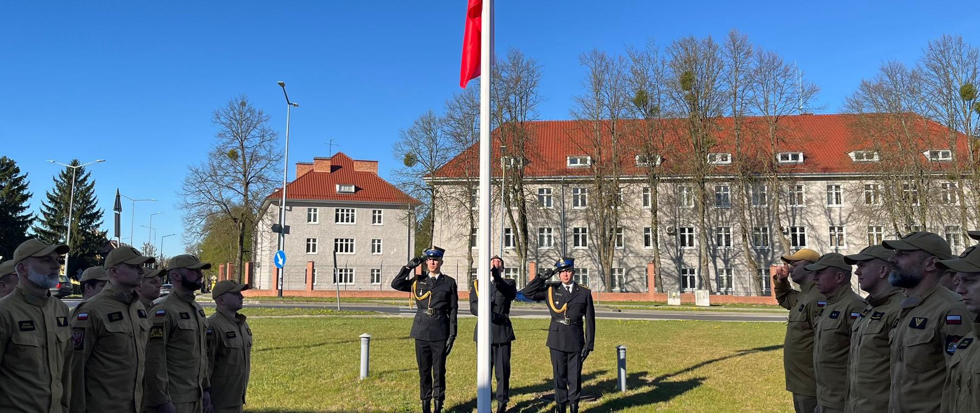 Uroczystość podniesienia polskiej flagi przed dużym budynkiem o czerwonym dachu. Dwóch funkcjonariuszy w ciemnych mundurach stoi przy maszcie i salutuje, a po obu stronach ustawieni są w równych szeregach strażacy w jasnych mundurach, również salutujący.