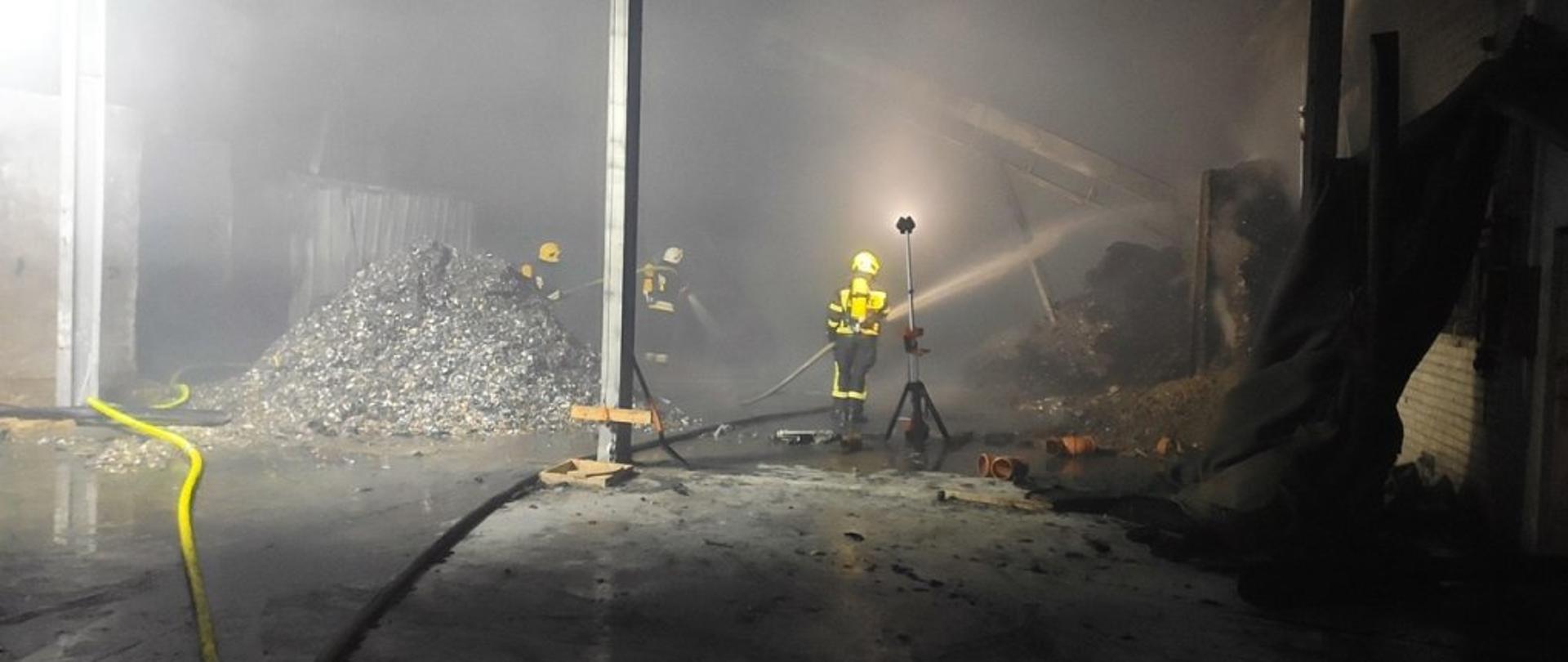 Zdjęcie zrobione wewnątrz częściowo spalonej hali, w której znajdują się śmieci. Na zdjęciu trzech strażaków w aparatach powietrznych gasi tlące się śmieci. W hali panuje duże zadymienie. Strażacy do oświetlenia miejsca pożaru korzystają z przenośnego zestawy oświetleniowego. 