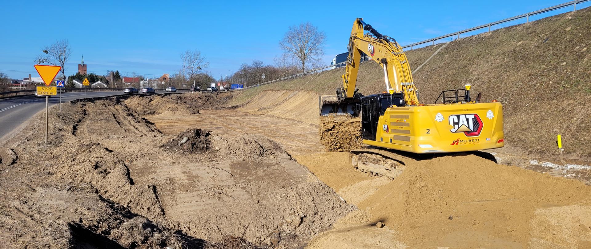 Zdjęcie przedstawia dużą koparkę gąsienicową pracującą na nasypie ziemnym wzdłuż drogi. Maszyna zrywa i przenosi warstwę piasku, tworząc szerokie wykopy i wyprofilowane skarpy. Po lewej stronie kadru widoczna jest jezdnia z ustawionymi znakami drogowymi, a w tle zabudowania miejscowości i drzewa. Niebo jest bezchmurne i intensywnie niebieskie.