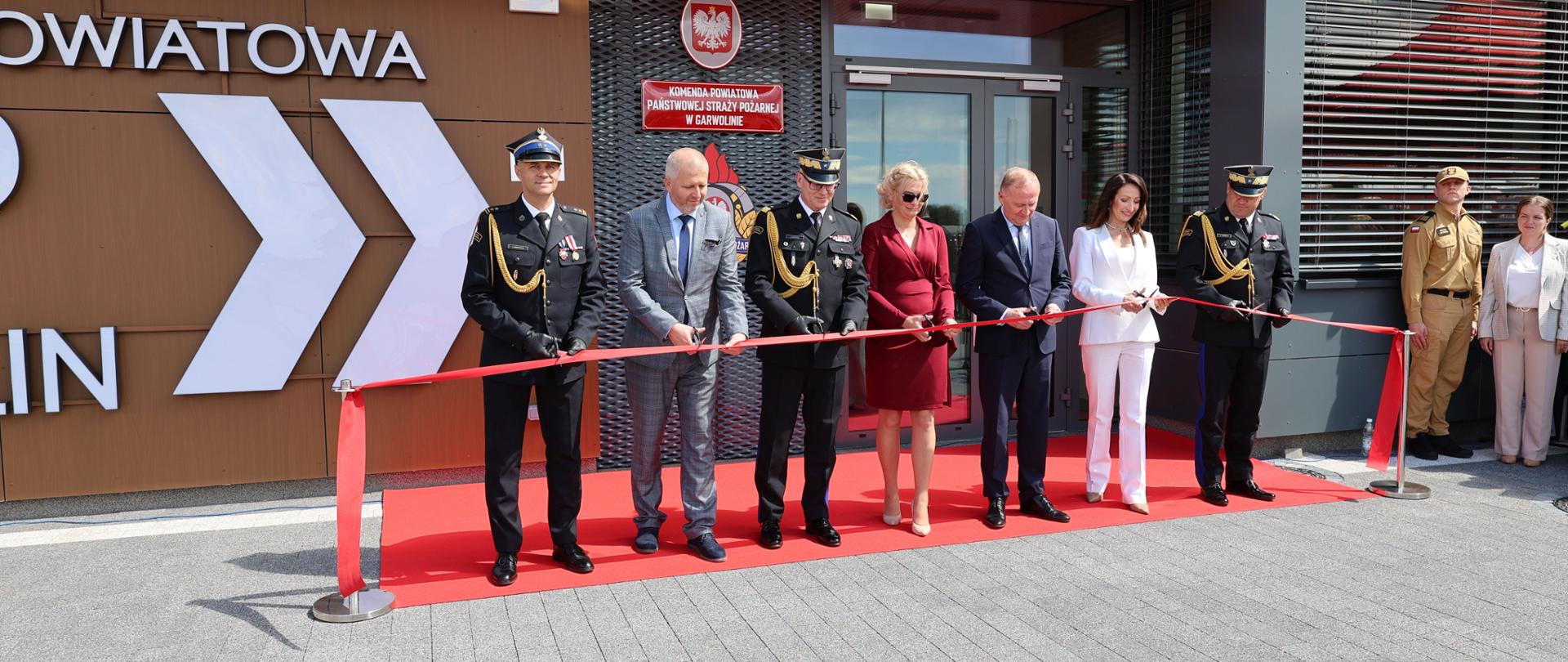 Grupa oficjeli oraz przedstawicieli straży pożarnej wspólnie przecina uroczystą czerwoną wstęgę na czerwonym dywanie. Ceremonia otwarcia ma miejsce przed wejściem do budynku Komendy Powiatowej Państwowej Straży Pożarnej w Garwolinie.