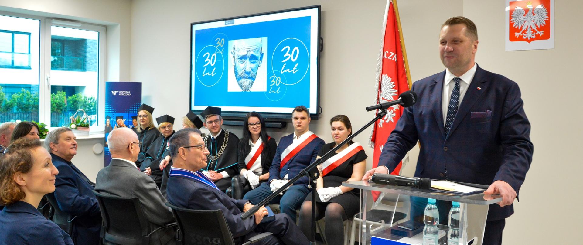 Minister Czarnek stoi za przezroczystą mównicą z tabliczką z napisem Uczelnia Korczaka i mówi do mikrofonu na stojaku, obok niego na ustawionych w rzędy krzesłach siedzi kilkanaście osób, w tle na białej ścianie wisi duży monitor, z lewej okna.