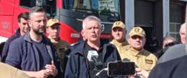 Na zdjęciu przedstawiono briefing prasowy z udziałem przedstawicieli Państwowej Straży Pożarnej oraz innych służb zaangażowanych w działania ratownicze. W centrum kadru znajduje się osoba udzielająca wypowiedzi mediom, przed którą ustawiono kamerę oraz mikrofony reporterskie. W tle widoczny jest samochód ratowniczo-gaśniczy PSP oraz funkcjonariusze w umundurowaniu służbowym i specjalnym. Fotografia została wykonana podczas przekazywania informacji dotyczących prowadzonych działań lub sytuacji kryzysowej.