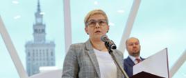 Pod półokrągłym oknem stoi wiceminister Mrówczyńska, mówi do mikrofonu i trzyma w ręku otwartą teczkę, za nią stoi dyrektor IBE.