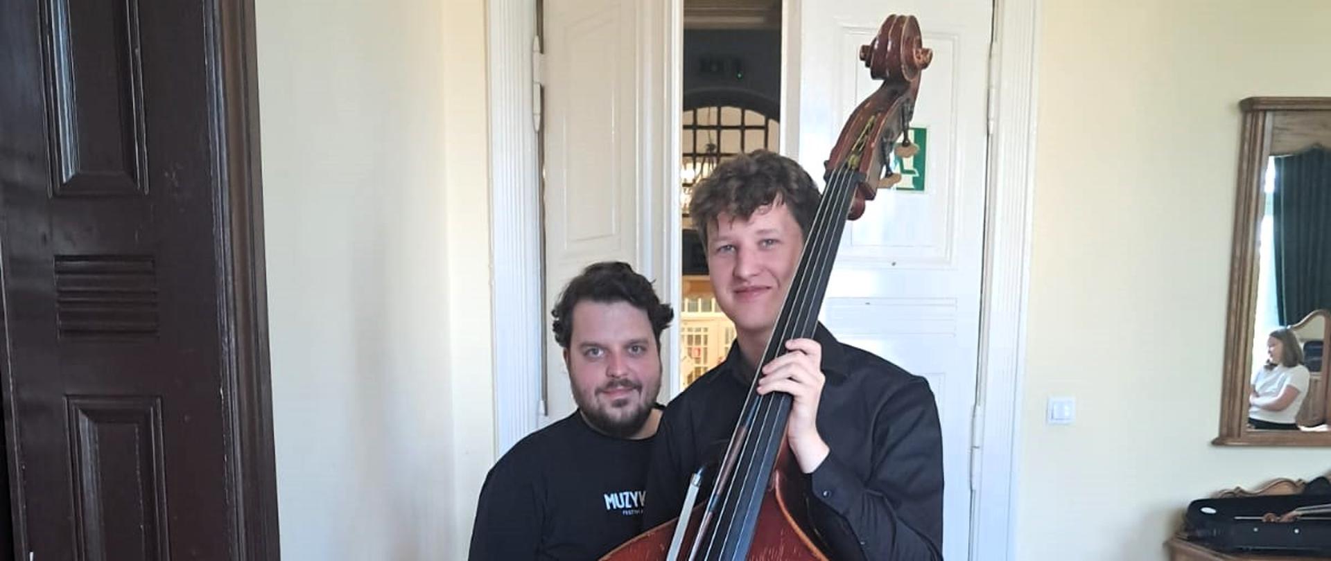 Dwóch mężczyzn uśmiecha się do zdjęcia. Pierwszy z nich, młodszy trzyma przed sobą kontrabas. Mężczyzna za nim jest ubrany na czarno i ma brodę.